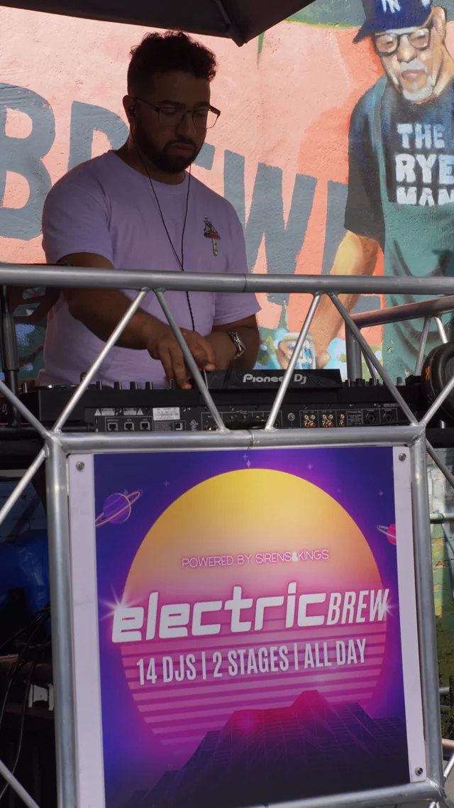 An Electric Brew DJ performing at an outdoor booth in daylight